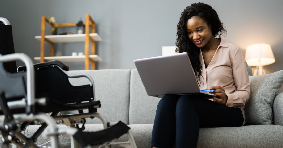 A woman sitting on a gray sofa using a laptop and credit card, with a wheelchair nearby inside a living room. A woman sitting on a gray sofa using a laptop and credit card, with a wheelchair nearby inside a living room.