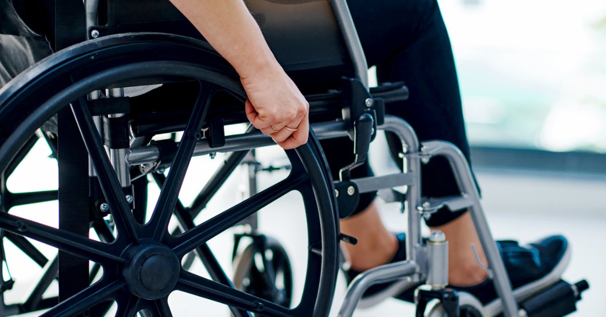 How to Build a Custom Setup With Wheelchair Parts A cropped person sitting in a wheelchair wearing dark pants and athletic black shoes. They are in a bright room.