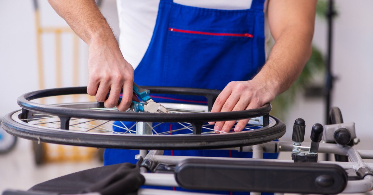 A person in overalls using pliers on a wheelchair wheel to repair it. The wheelchair has a metal frame. A person in overalls using pliers on a wheelchair wheel to repair it. The wheelchair has a metal frame.