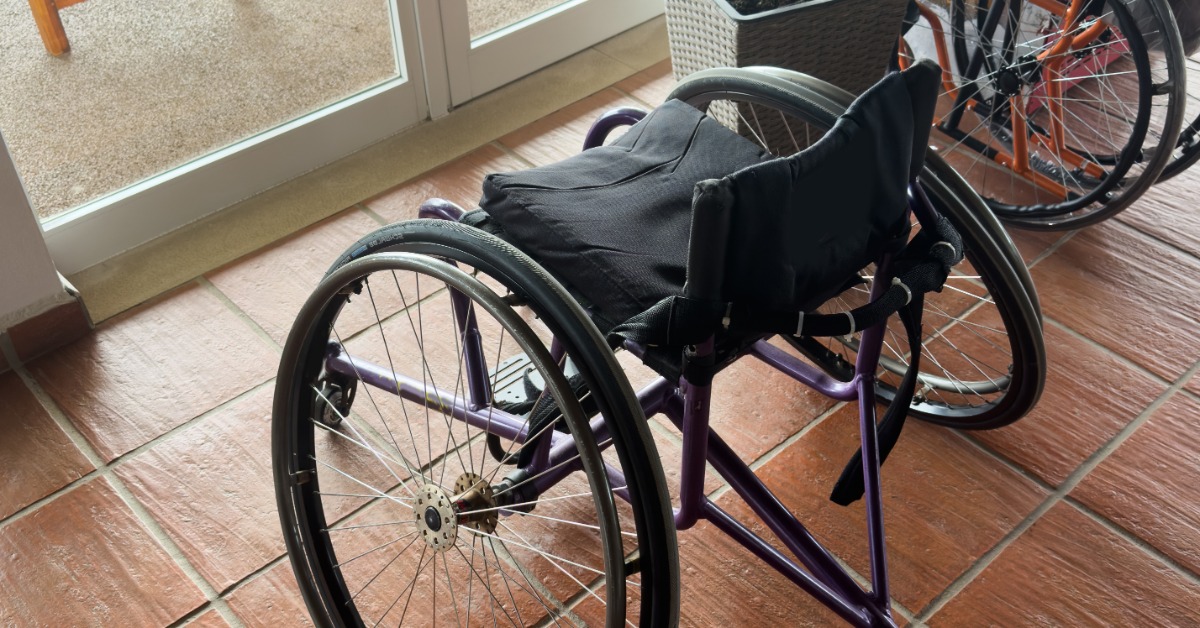 A wheelchair with a purple frame and black cushioned seat resting on tile flooring near glass doors.