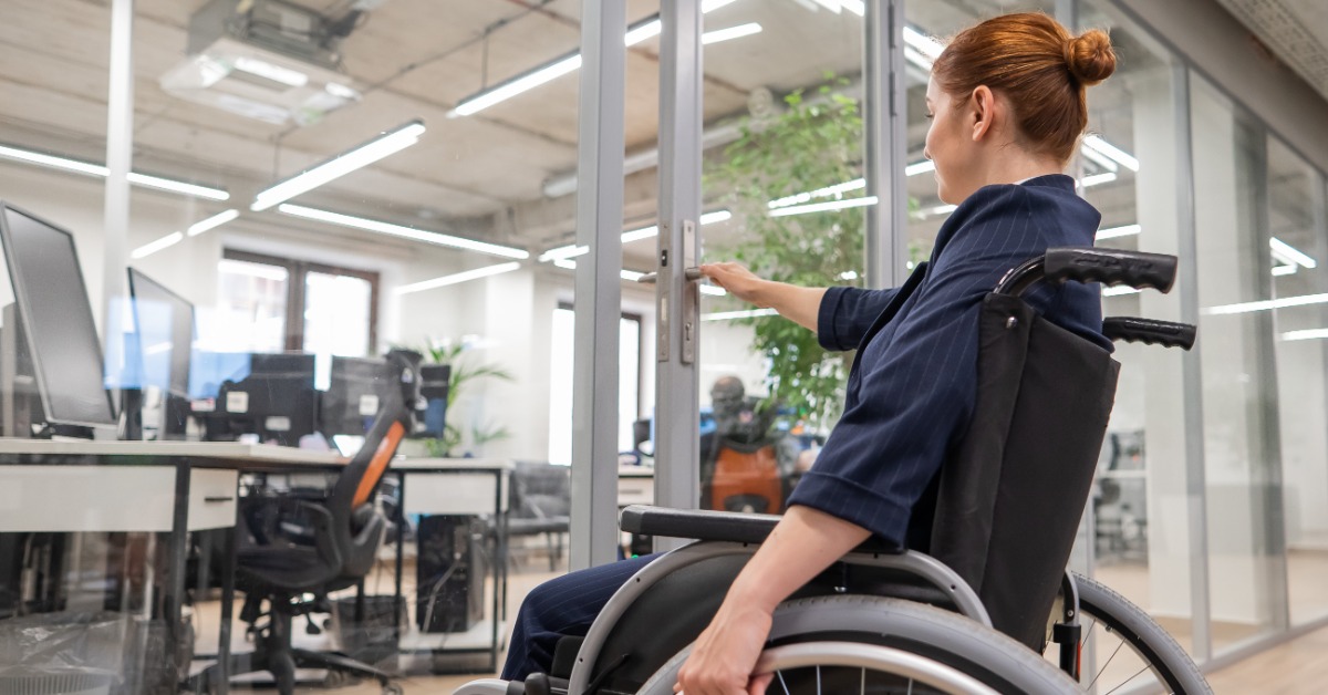 A red-haired woman seated in a wheelchair reaching for a glass office door handle, with desks and chairs behind it. A red-haired woman seated in a wheelchair reaching for a glass office door handle, with desks and chairs behind it.