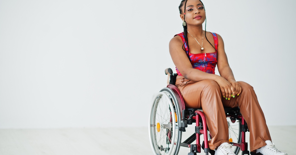 A young woman wearing a red and blue top, white sneakers, and brown pants sits in a black wheelchair.