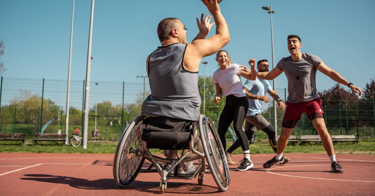 A man in a wheelchair on a park court shoots a basketball. Three other able-bodied people are also playing.