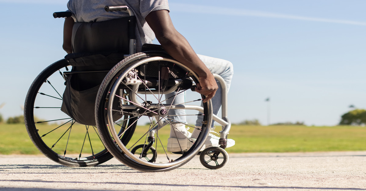 A man wearing a T-shirt and jeans in a manual wheelchair in the park on a sunny day facing away from the viewer.