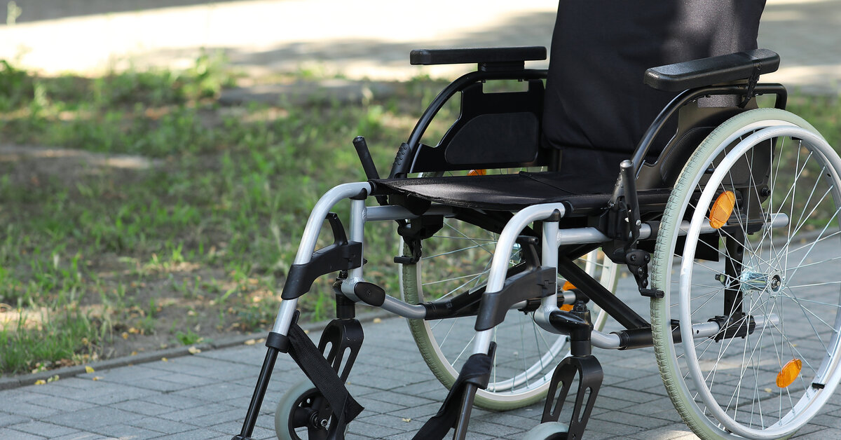 An empty manual wheelchair with a black seat and backrest sits outside on a paver path near grass in the shade.