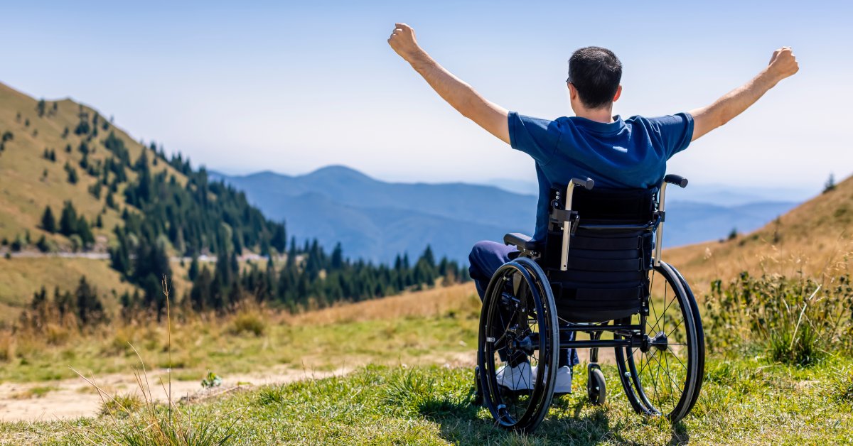 A man wearing a blue t-shirt sitting in a wheelchair faces a scenic mountainside, raising his arms in accomplishment.