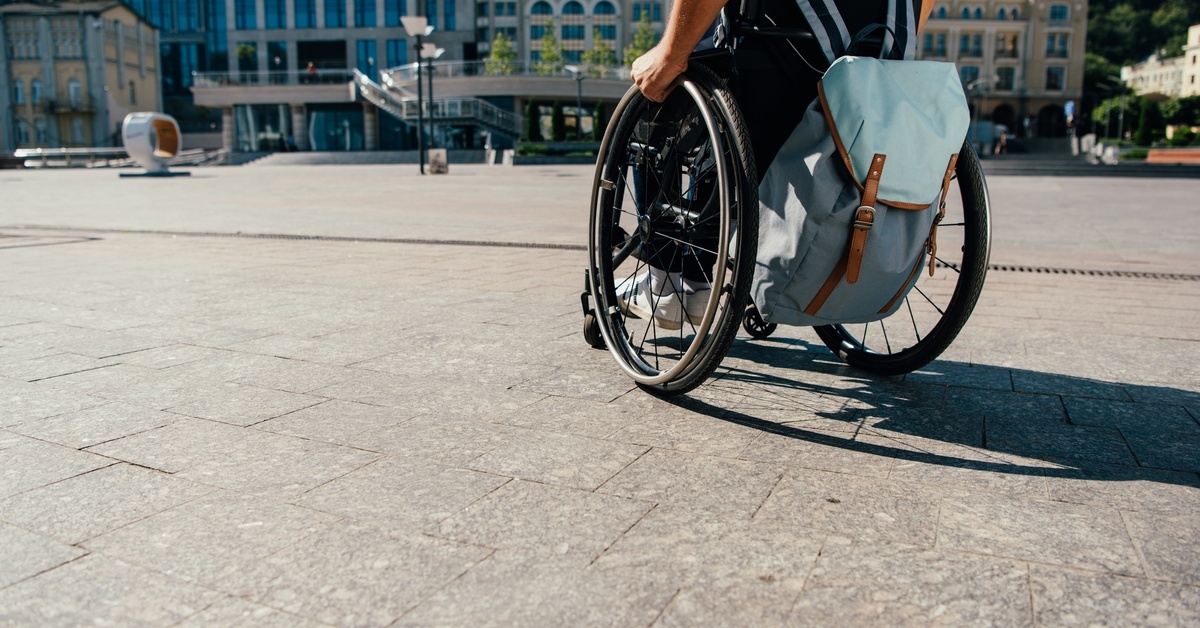 A man in a wheelchair with a light blue backpack on the chair pushes himself through a city plaza on a sunny day.