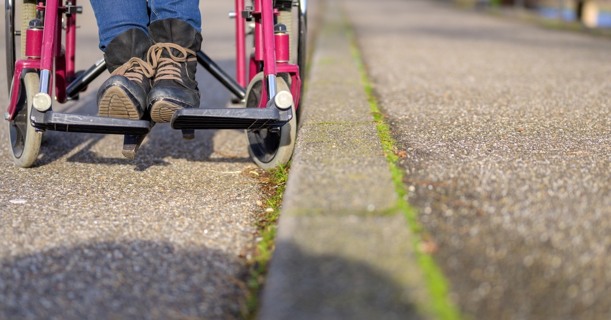 A low-angle view of a woman wearing heeled boots and jeans in a manual wheelchair with a pink frame against a curb.
