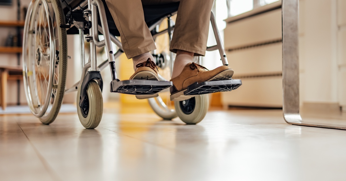 A low-angle view of a man, wearing beige pants and loafers, riding in a wheelchair indoors with a blurred background.