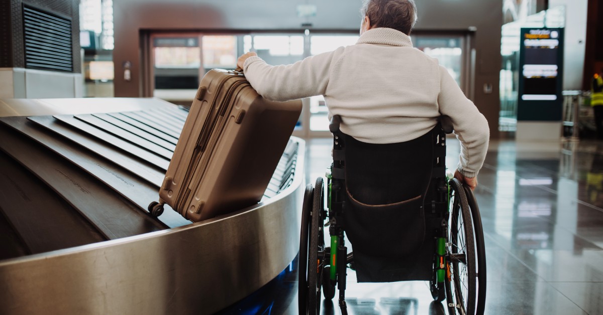 The backside of a man sitting in a wheelchair. He has his hand on the handle of a suitcase that is on a conveyor belt.