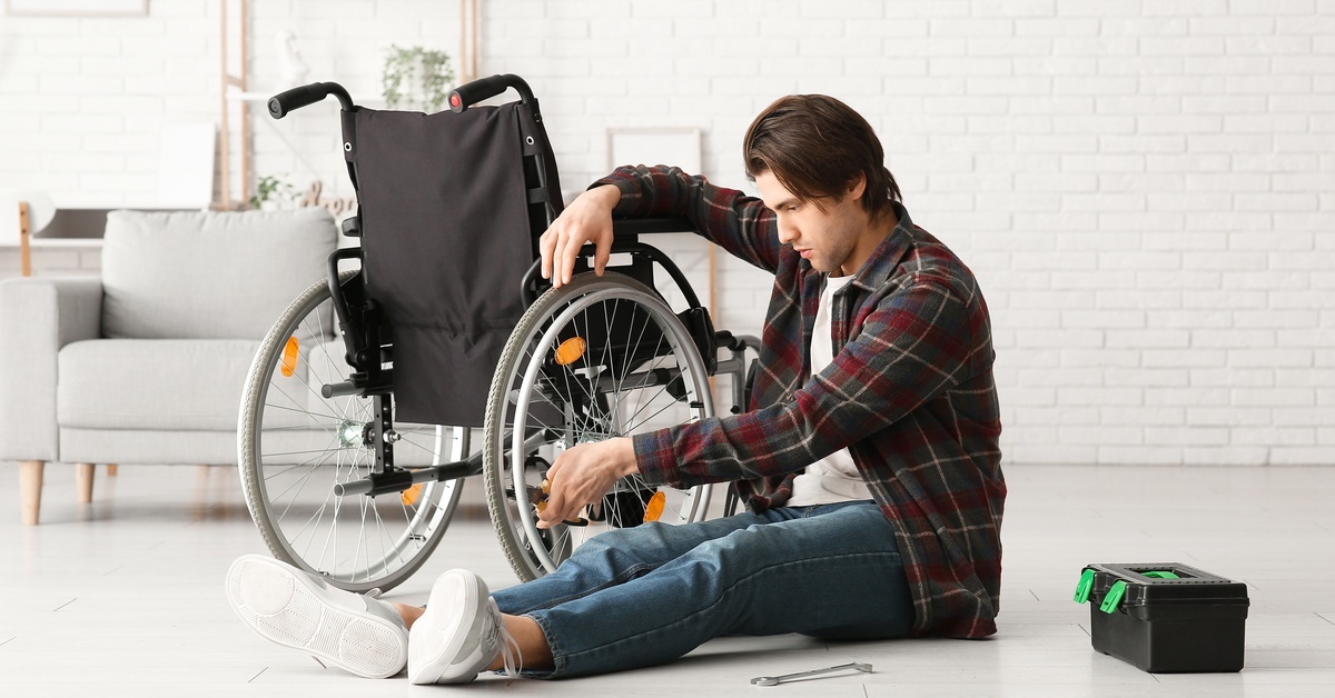 A young man sitting on the floor next to his wheelchair. He is using a pair of pliers to repair the wheel.