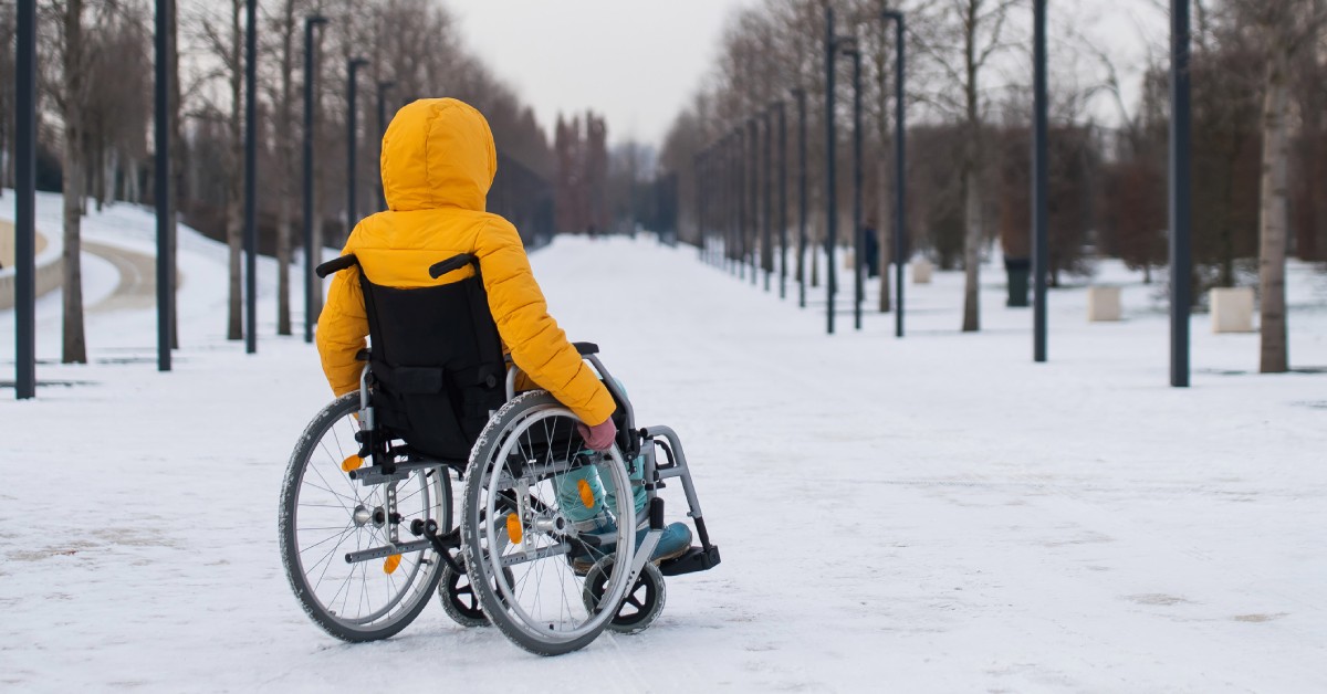 A woman wearing a winter coat sitting in a wheelchair outside. There is snow on the ground and bare trees around her.