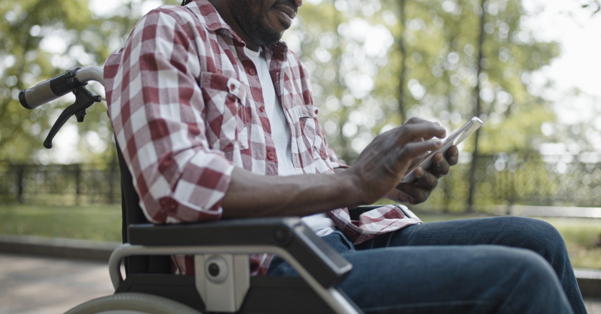A man in a wheelchair sitting in an outdoor area. He is looking down at his phone to make an online order.