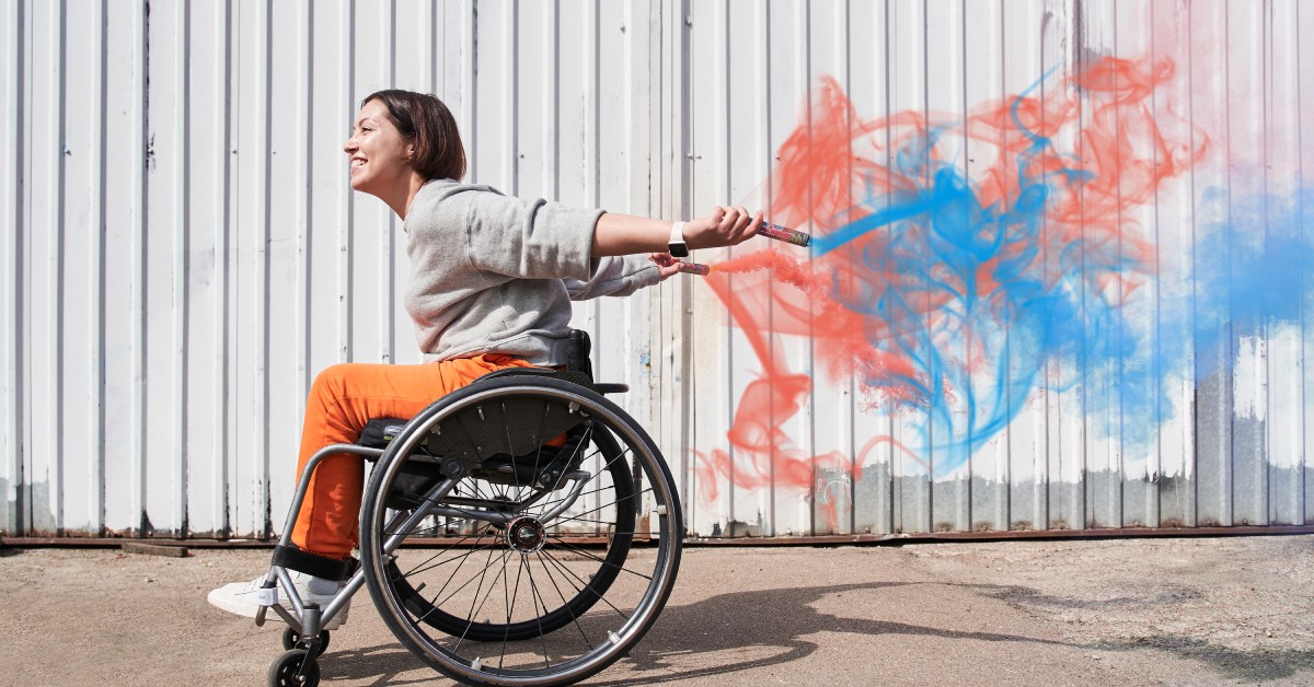 A young woman in a wheelchair holding two smoke wands that are emitting blue and red smoke behind her.