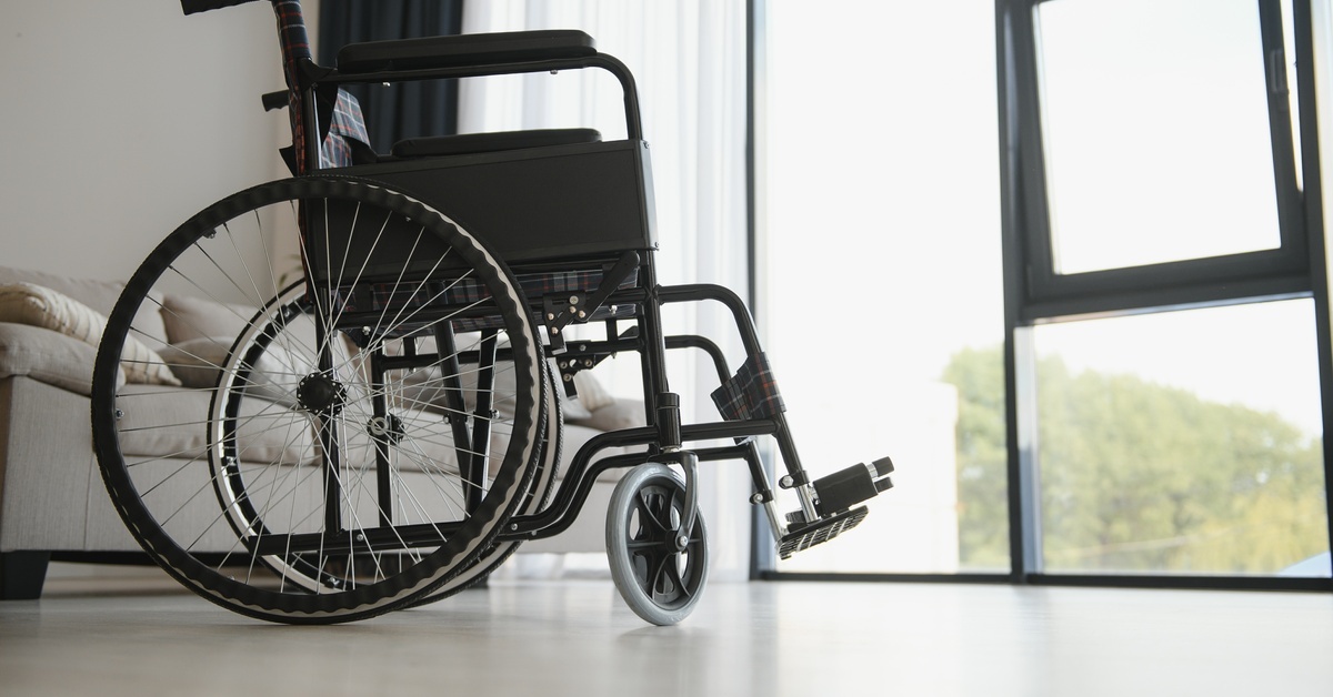 An empty wheelchair in a well-lit living room that has a couch and a large window showing trees outside.