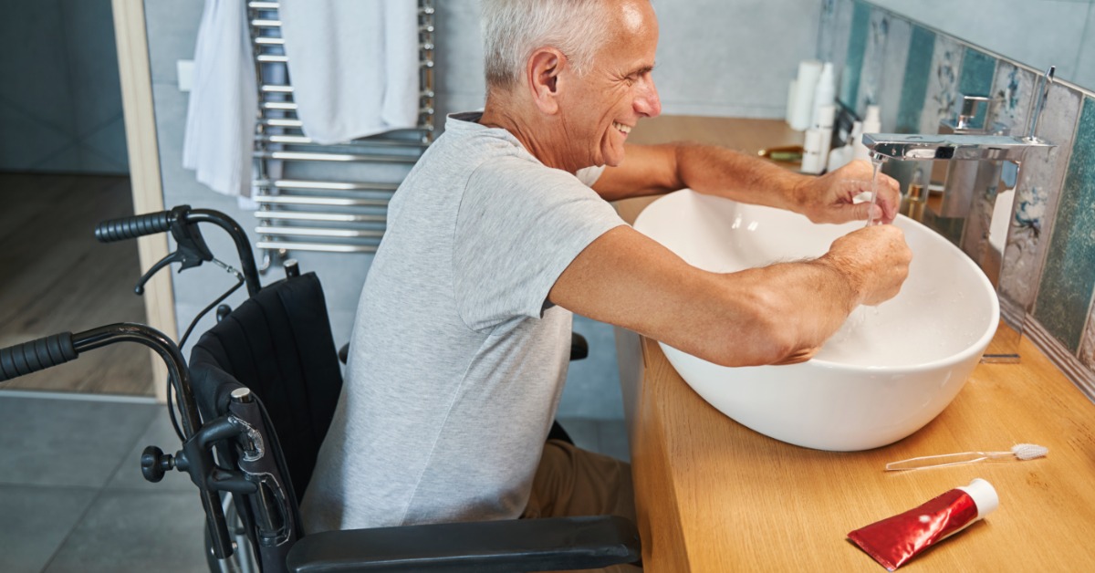 A man in a wheelchair washing his hands at an accessible sink in the bathroom. His wheelchair fits under the sink.