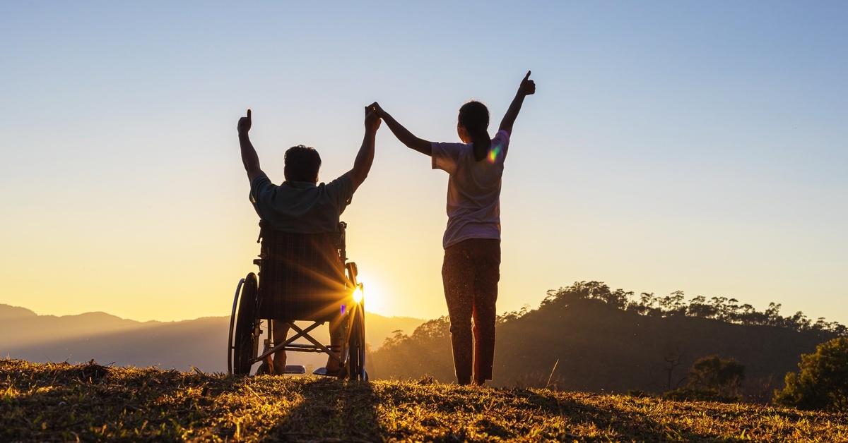 Two people on a hill with their arms up, looking at the sunset. One of the individuals is in a wheelchair.