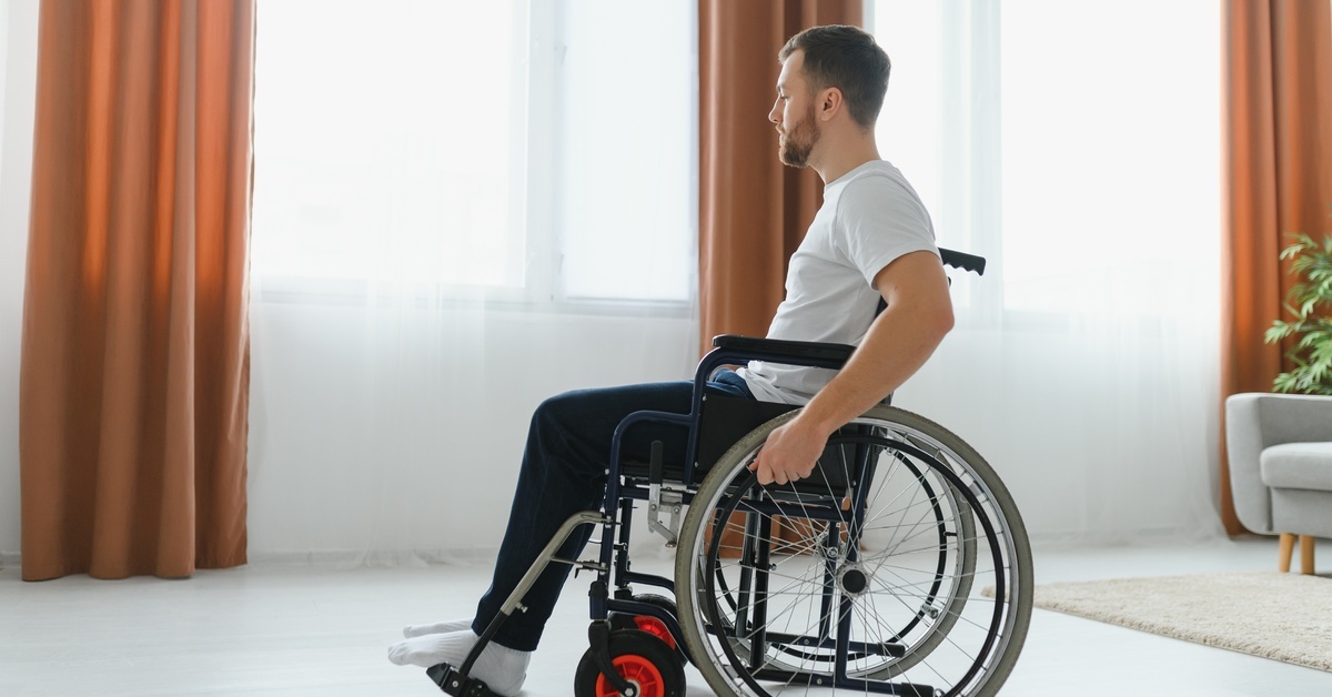 A young man in a wheelchair with spoke wheels. He is in a brightly lit living room, sitting in front of the windows.