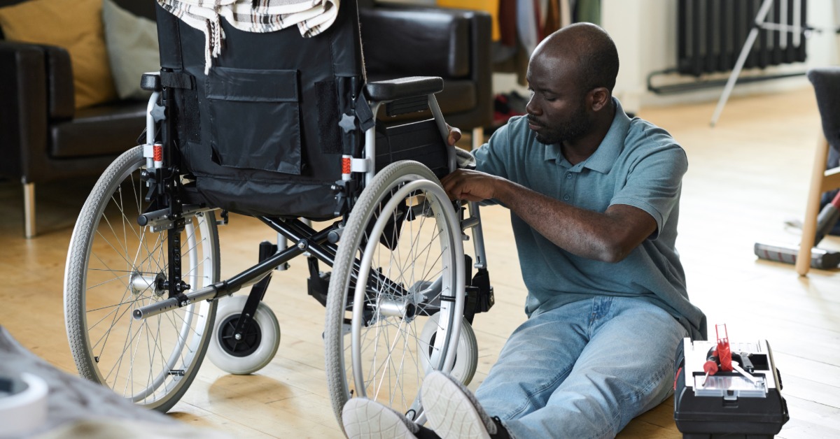 A man with a disability sitting on the floor, working on repairing his wheelchair. There is a repair box next to him.