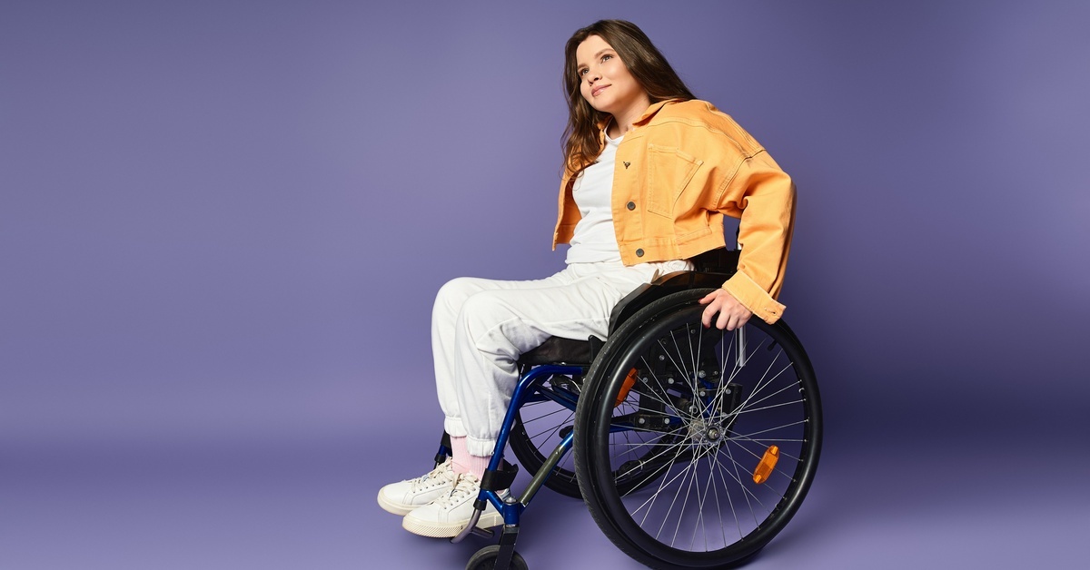A young woman in a yellow jacket sitting in a wheelchair. She has her hands on the wheel to move herself.