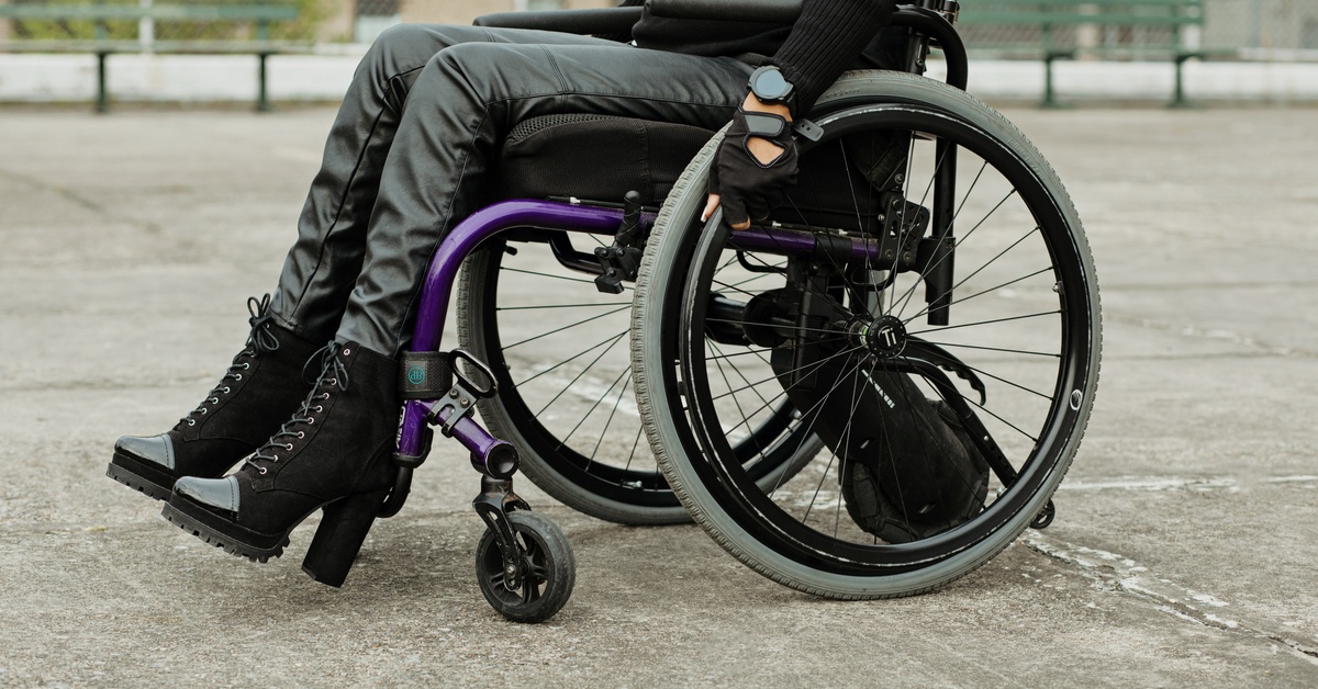 A person wearing black boots and gloves sitting in a purple wheelchair on a concrete surface outdoors.