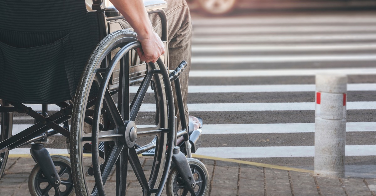 A disabled man in a wheelchair waiting at a crosswalk to go across the road. He has his hand on the wheel to move.