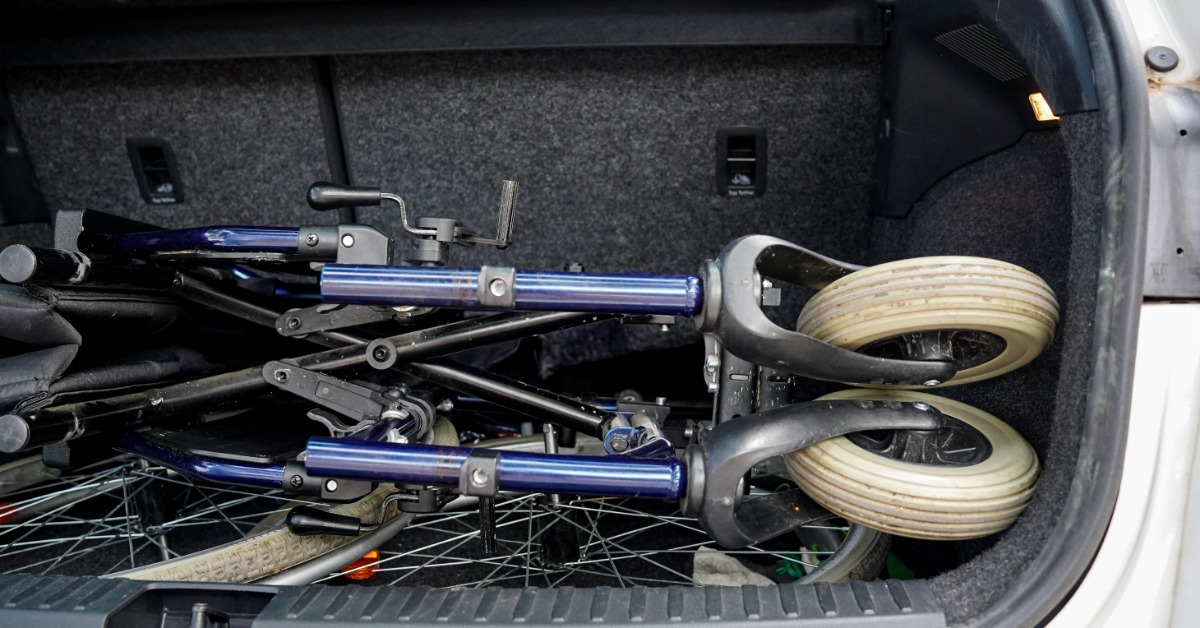 A folded wheelchair lying in a car trunk with gray upholstery. The wheelchair has blue legs and slightly dirty wheels. A folded wheelchair lying in a car trunk with gray upholstery. The wheelchair has blue legs and slightly dirty wheels.